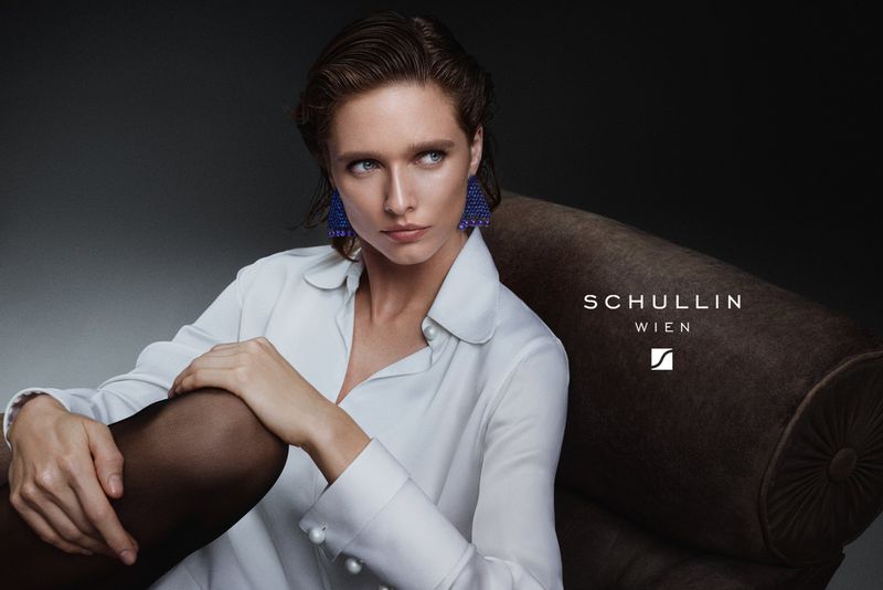 Elegant woman in a white blouse and blue earrings sitting on a sofa in a photo studio, looking off camera with a neutral expression.