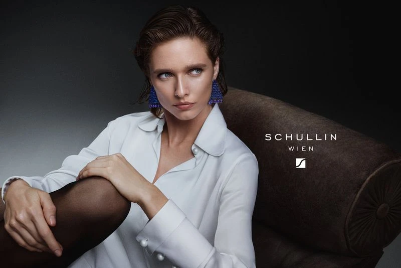 Elegant woman in a white blouse and blue earrings sitting on a sofa in a photo studio, looking off camera with a neutral expression.