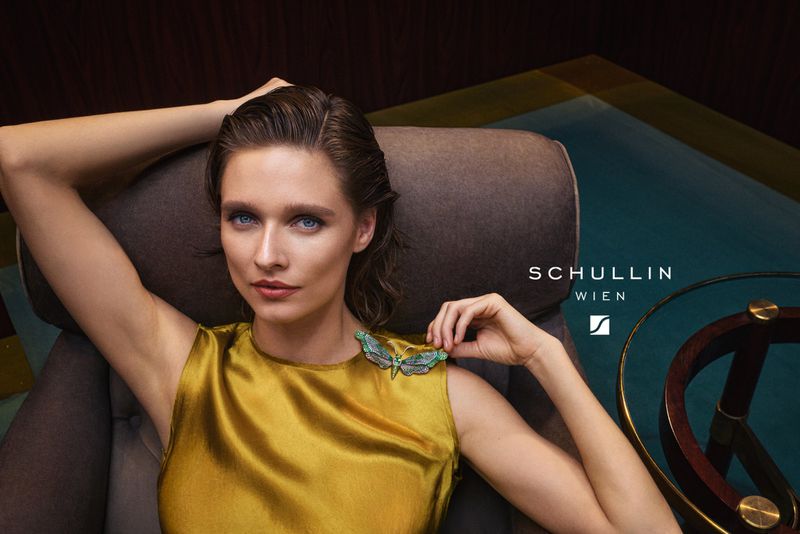 A woman with fair skin poses in a gold satin dress, seated in a chair with an olive-green butterfly brooch pinned to her shoulder.