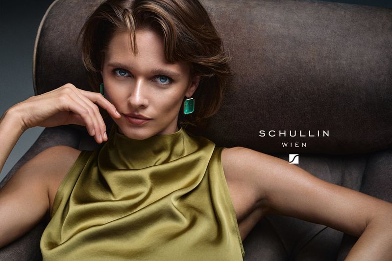 Close-up of a young woman with olive-toned skin wearing a sleeveless green silk top and emerald earrings, sitting on a brown chair.