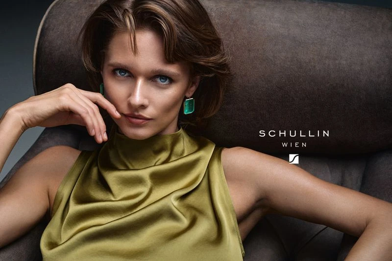 Close-up of a young woman with olive-toned skin wearing a sleeveless green silk top and emerald earrings, sitting on a brown chair.