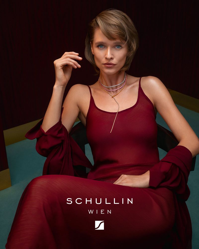 Elegant woman in a red slip dress and jewelry sitting indoors in a chair; jewelry advertisement.