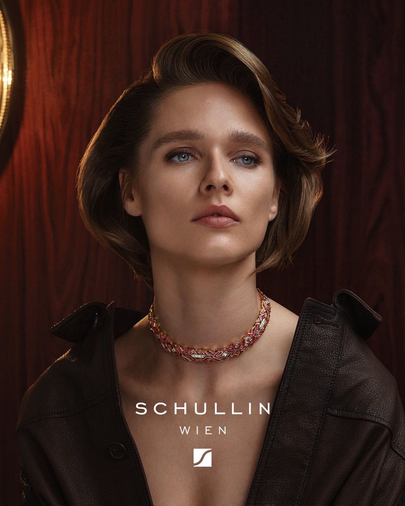 Close-up portrait of a woman wearing a jeweled choker necklace and dark jacket against a dark wood background for Schullin Wien.