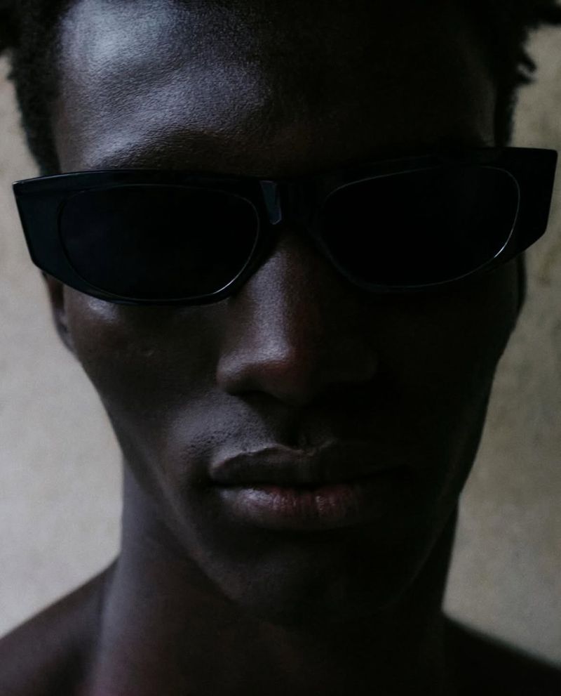 Close-up portrait of a dark-skinned adult male wearing black sunglasses, looking directly at the camera against a neutral background.