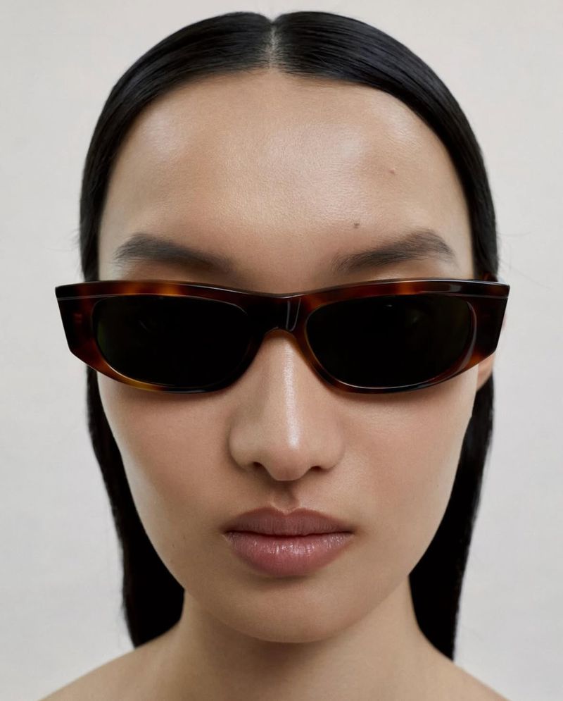 Close-up portrait of a woman with dark hair wearing tortoiseshell sunglasses against a plain white background in studio lighting.