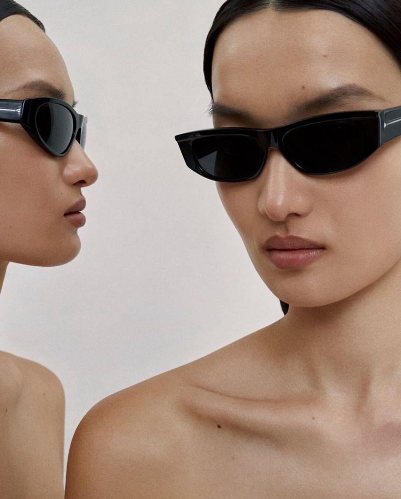 Close-up portrait of a model wearing dark black sunglasses against a clean neutral background for Oliver Peoples.