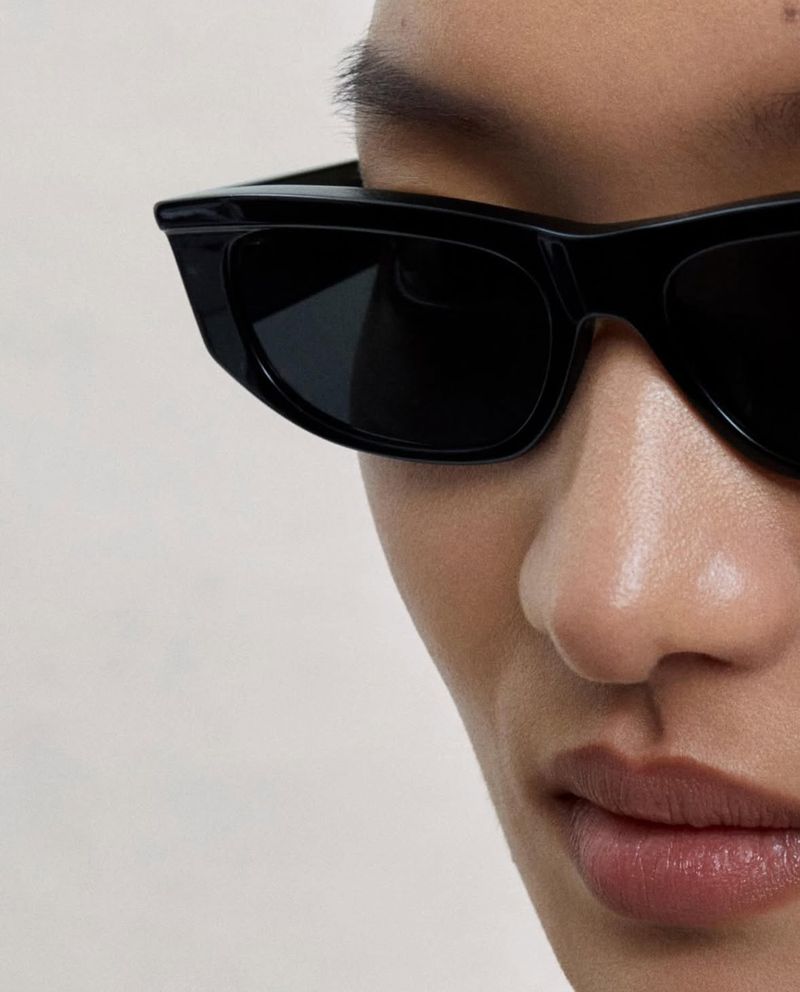 Close-up portrait of a model wearing black sunglasses against a neutral background for Oliver Peoples eyewear.