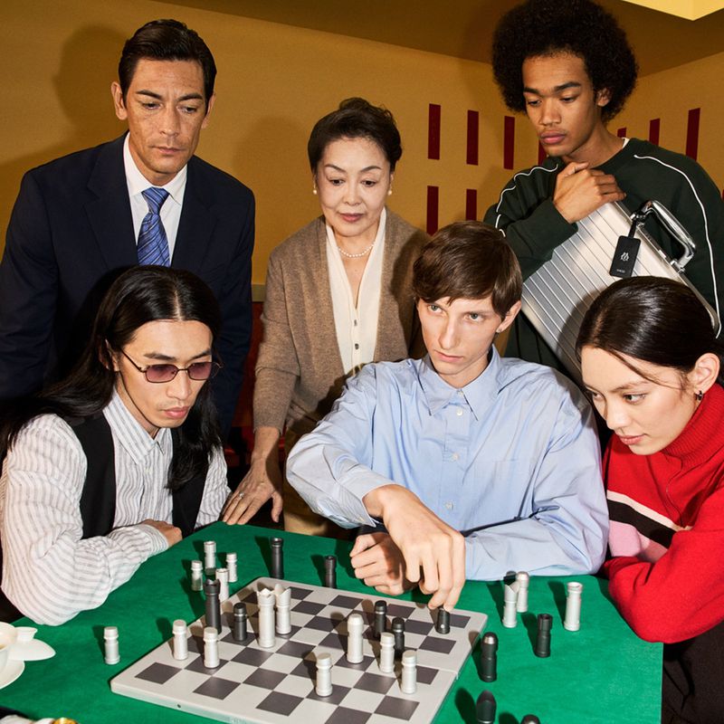 Group of six people gathered around a chessboard in an indoor setting with cream and red wall details, creating a contemplative atmosphere.