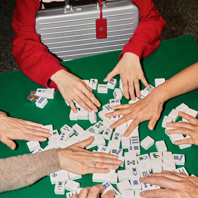 Overhead shot of multiple adults playing Mahjong with tiles arranged on a green table, along with hands, arms, and a silver briefcase.