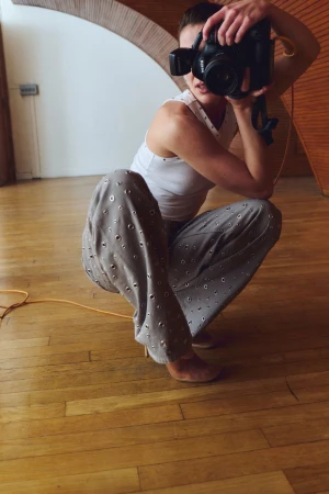 Woman squats on a wood floor, holding up a camera. The room features arched doorway, and neutral wall colours with natural light.