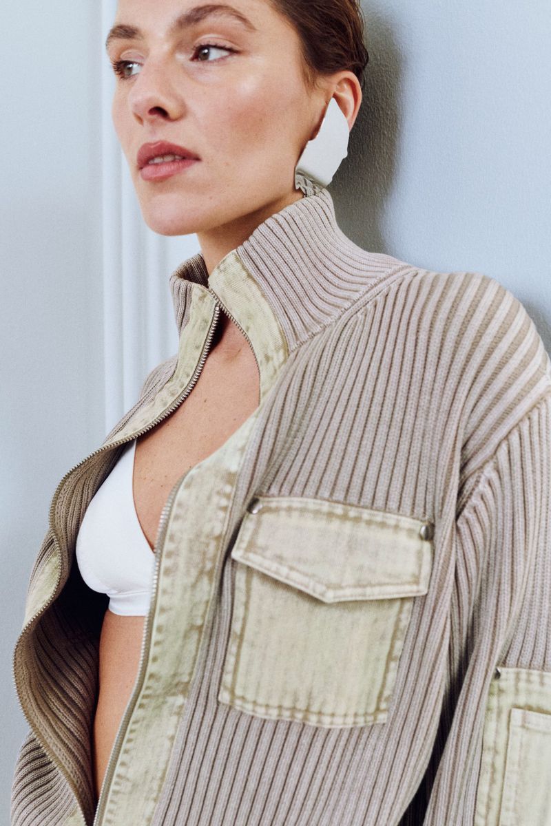 A woman wears a white bra and beige zip-up jacket with large earrings, posing indoors against a light-colored wall in a medium shot.