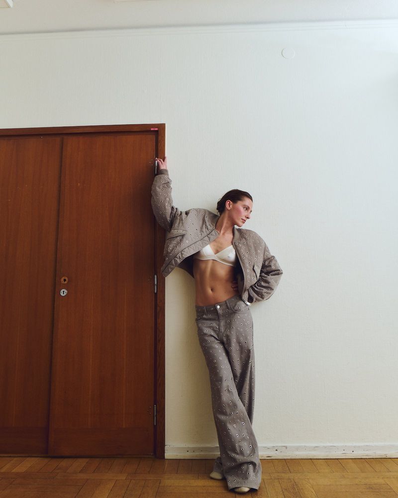 Model leaning against a wooden door wearing an embellished grey jacket and matching wide-leg trousers for Weekday.