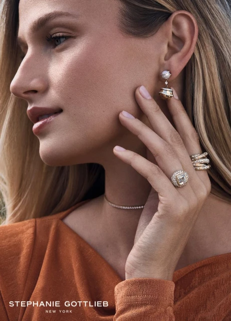 Close-up studio shot of a fair-skinned blonde woman modeling diamond jewelry, including earrings, a ring, and a necklace, on a neutral background.