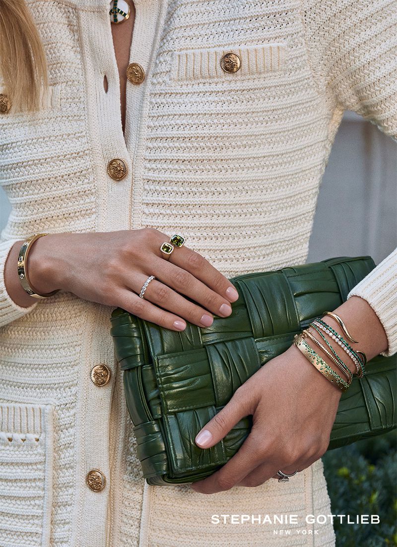 Close-up of a woman wearing diamond rings and gold bracelets holding a woven green leather handbag against a cream knit outfit.