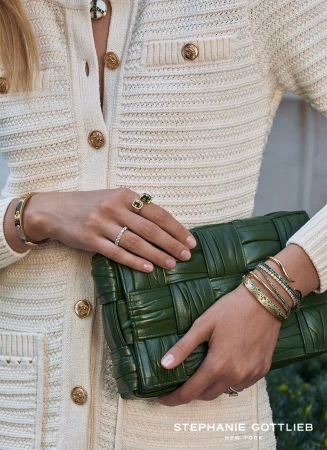 Close-up of a woman wearing diamond rings and gold bracelets holding a woven green leather handbag against a cream knit outfit.