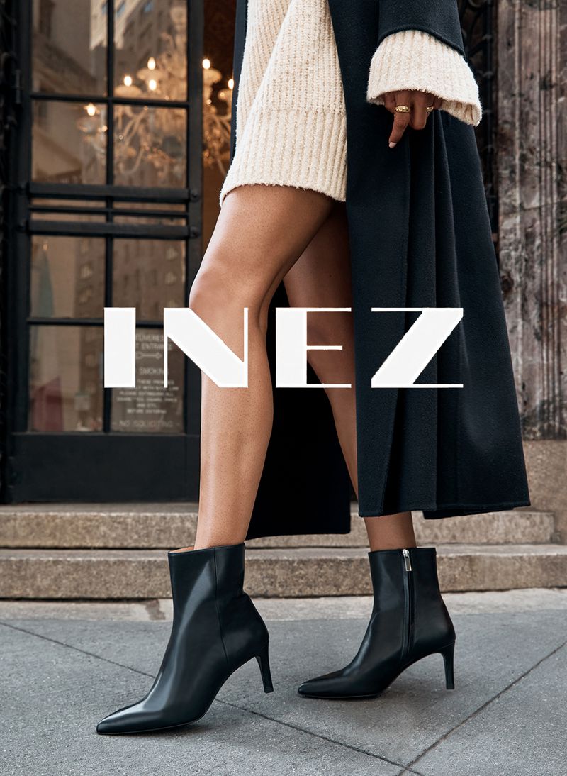Fashion shot: woman wearing black ankle boots and a cream sweater dress, standing on a sidewalk with a building in the background.