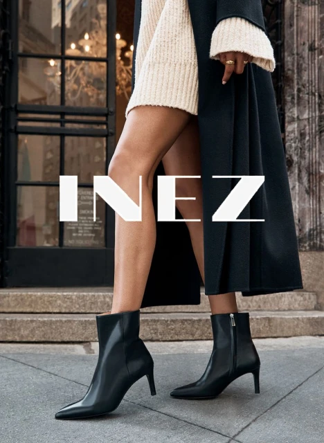 Fashion shot: woman wearing black ankle boots and a cream sweater dress, standing on a sidewalk with a building in the background.
