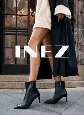 Fashion shot: woman wearing black ankle boots and a cream sweater dress, standing on a sidewalk with a building in the background.