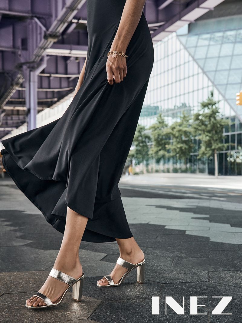 Woman in a black dress and silver high-heeled sandals walking on a city street with buildings and an elevated train overpass.