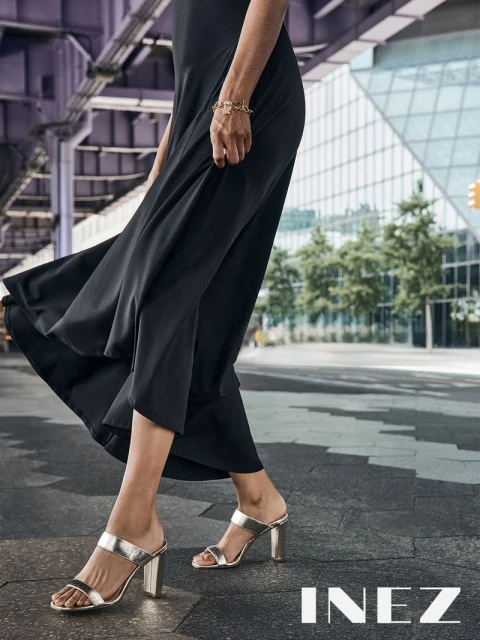 Woman in a black dress and silver high-heeled sandals walking on a city street with buildings and an elevated train overpass.