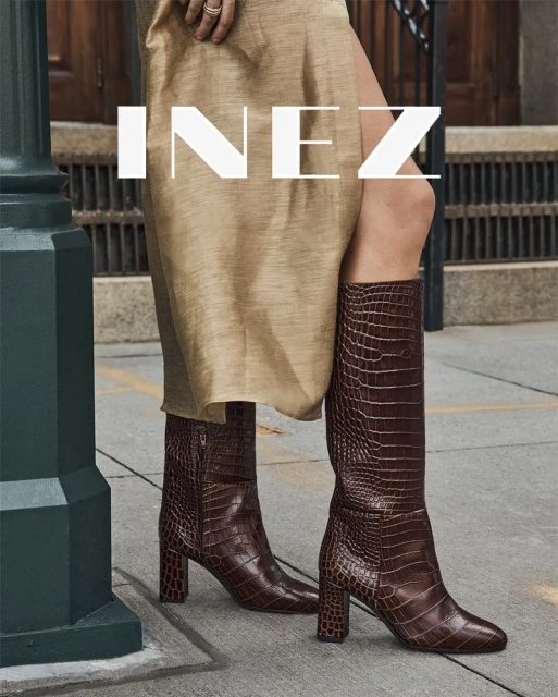 Woman in brown reptile-print knee-high boots stands on an urban sidewalk next to a green lamppost and wears a beige dress.