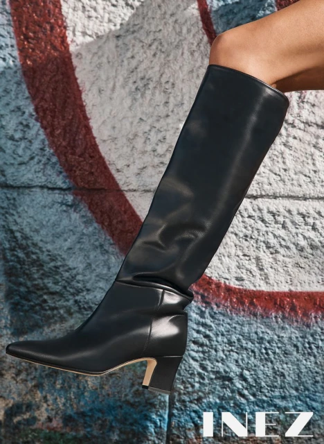 Black leather knee-high boot on a model, showcased against a graffiti-style painted wall, creating a street-meets-high-fashion aesthetic.