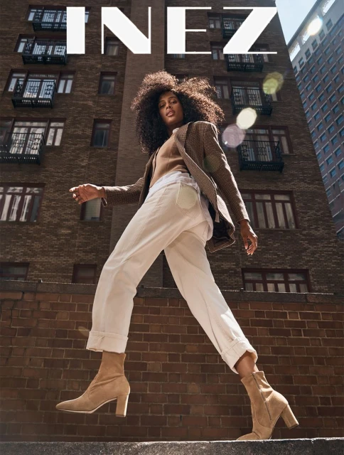 Fashion editorial shot, low-angle, woman walking confidently in blazer, tan top, cuffed pants, and heeled booties in an urban setting.