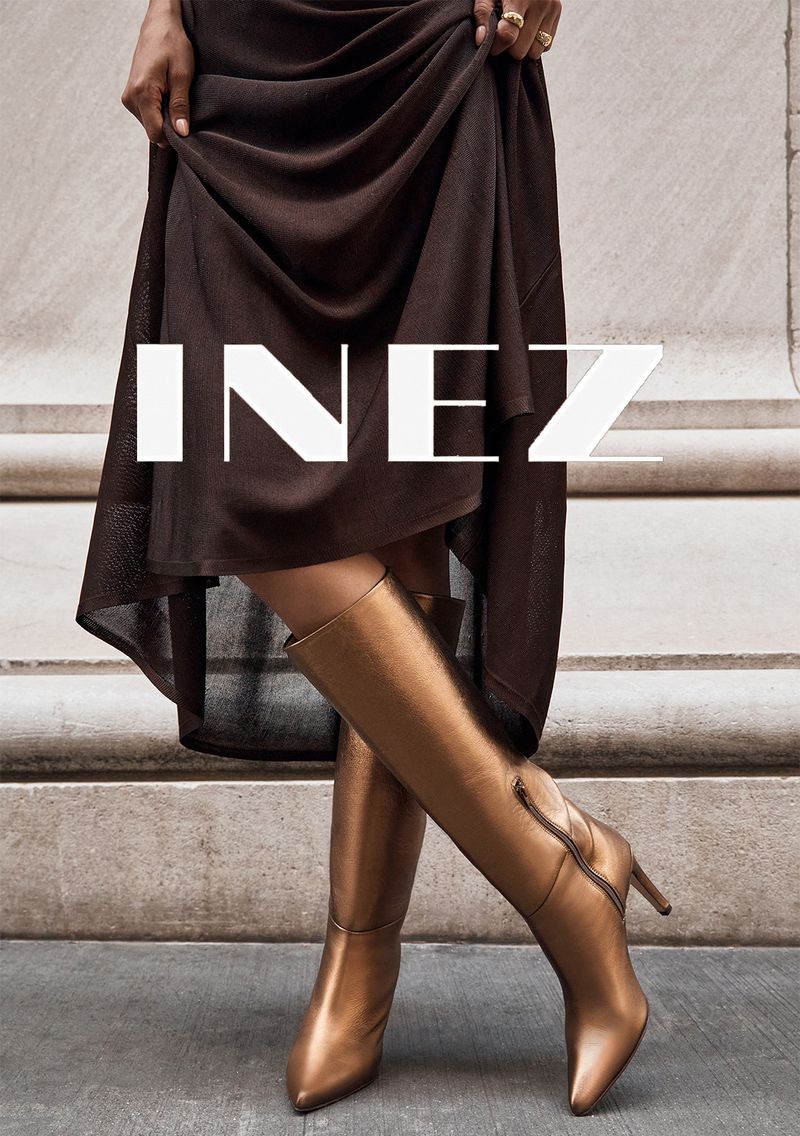 Elegant bronze knee-high heeled boots paired with a flowing brown dress and a stylish gold ring, against a neutral architectural backdrop.