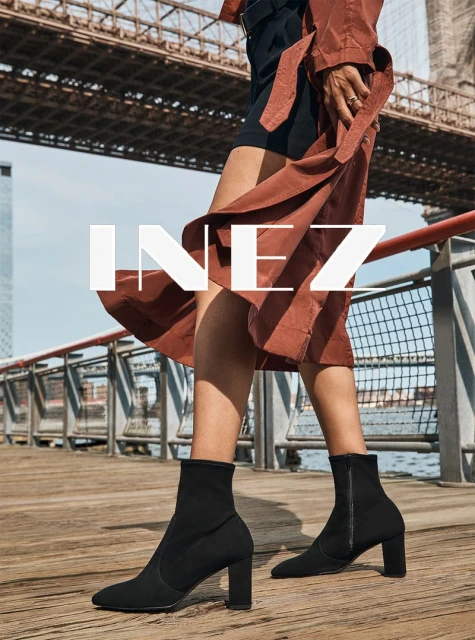 Woman wearing a brown trench coat and black ankle boots walking on a wooden boardwalk with the Brooklyn Bridge in the background.
