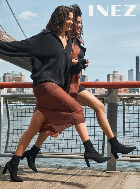Two adult women walk on a city boardwalk next to a bridge wearing sweaters, skirts, and ankle boots in daytime.