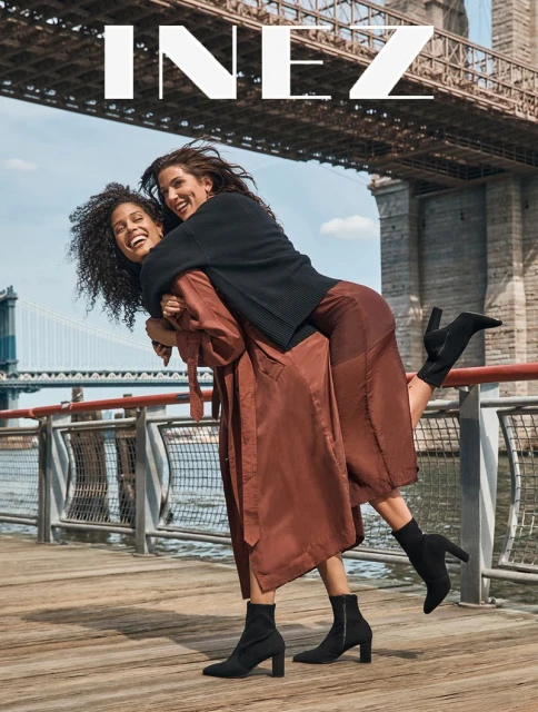 Two women laugh on a waterfront pier with a visible bridge in the background, modelling stylish black ankle boots and clothing.