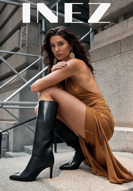 A woman poses on a concrete staircase in a tan dress and black boots, with architectural elements visible in the background.