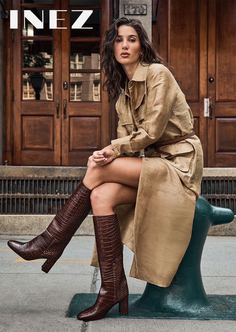 Adult woman in a tan trench coat and brown knee-high boots poses outside a brick building. Fashion editorial street style.