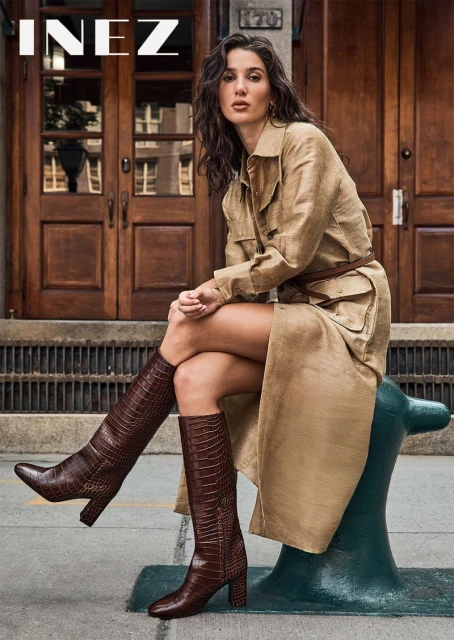 Adult woman in a tan trench coat and brown knee-high boots poses outside a brick building. Fashion editorial street style.
