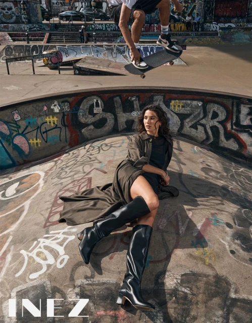 A woman in a coat and boots poses inside a skate park, while a skateboarder catches air in the background, creating an edgy fashion shot.