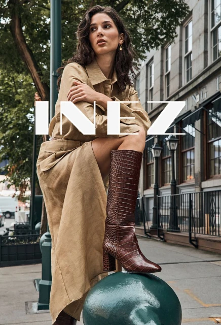 A woman in a trench coat and boots poses in an urban scene, showcasing a fashion-forward look with architectural details in the backdrop.