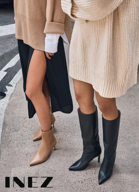 Two women stand on a city street, one in tall black leather boots, the other in tan ankle boots, both showcasing fall fashion.