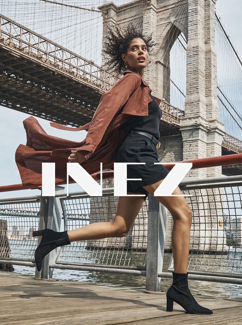 A woman in a brown coat and black boots runs across a wooden pier with the Brooklyn Bridge in the background, creating a dynamic fashion image.