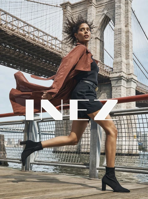 A woman in a brown coat and black boots runs across a wooden pier with the Brooklyn Bridge in the background, creating a dynamic fashion image.