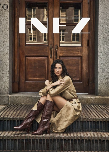A brunette woman in a tan trench coat and brown boots sits on concrete steps in front of a brown wooden door. Fashion editorial shot.