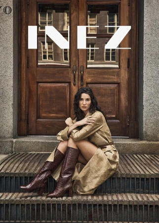 A brunette woman in a tan trench coat and brown boots sits on concrete steps in front of a brown wooden door. Fashion editorial shot.