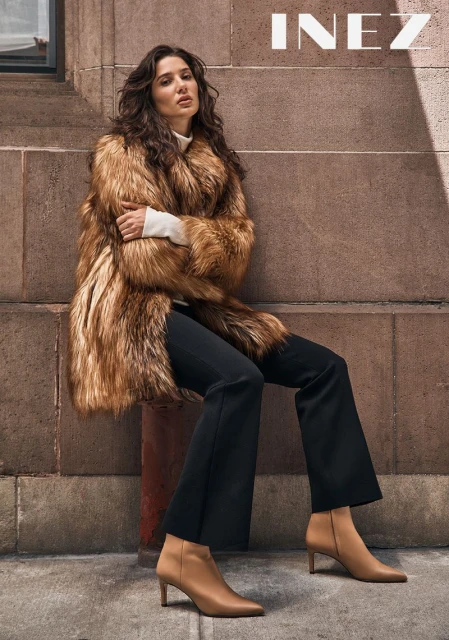 A fashionable adult woman in a fur coat, black pants, and tan boots poses against a stone wall and column in an outdoor setting.
