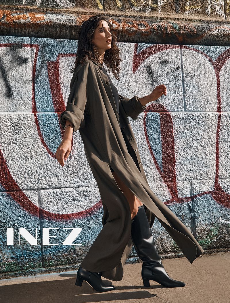 A brunette woman in a long green coat and black boots poses against a graffiti wall, showcasing fashion and urban style in natural light.