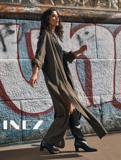 A brunette woman in a long green coat and black boots poses against a graffiti wall, showcasing fashion and urban style in natural light.