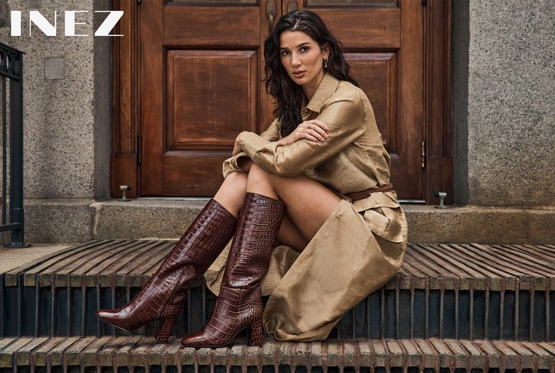 Woman in a beige trench coat seated on wooden steps wearing tall brown boots, photographed by Yossi Michaeli for Inez.