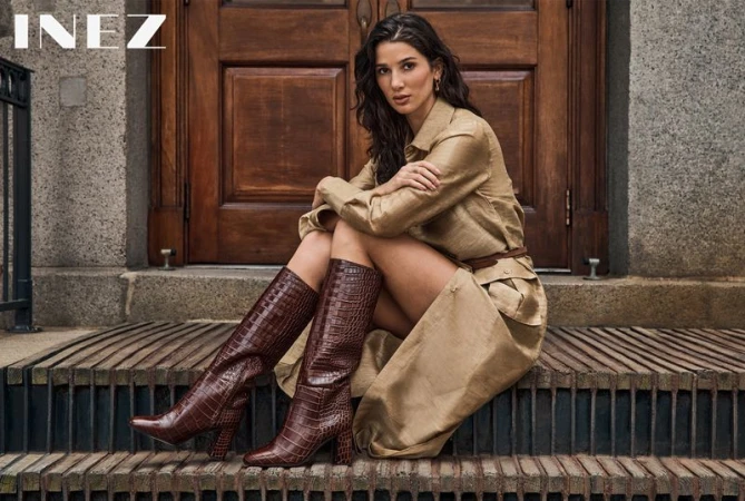 Woman in a beige trench coat seated on wooden steps wearing tall brown boots, photographed by Yossi Michaeli for Inez.