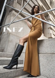 Model wearing a long gold dress and black knee-high boots posing outdoors on stone steps beneath architectural scaffolding.