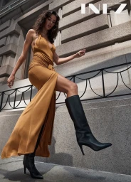 Model in a halter neck gold dress and black knee-high boots stepping over a railing in front of a stone building.