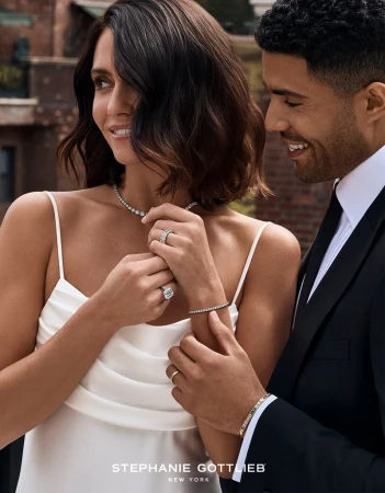 A woman in a white silk slip dress wearing diamond jewelry and a man in a black tuxedo smiling outdoors.