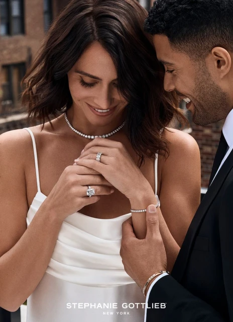 Woman and man showcasing diamond jewelry: necklace, rings, and bracelets, in front of a blurred cityscape on a sunny day.
